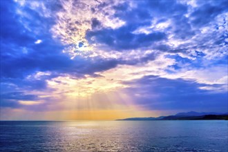 Beautiful seascape with colorful clouds and bright sun rays cast over calm sea waters and hazy distant shores. Sunrise or sunset in Mediterranean sea