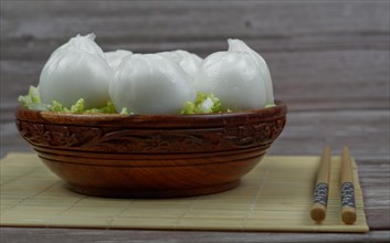 Xiao long bao of prawns on a bed of lettuce in a wooden bowl with chopsticks and a ceramic spoon with soy sauce