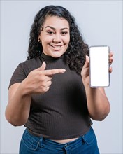 Smiling latin girl showing and pointing at cell phone screen isolated. Funny girl with curly hair showing an advertisement on her cell phone