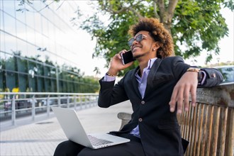 Business person talking to the mobile phone and smiling while working on a park bench