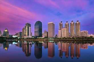 Beautiful landscape with modern high-rise buildings reflecting in calm lake waters by park and promenade at colorful sunset. Benjakitti Park