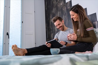 Side view of a happy couple spending time on bed with a digital tablet and coffee cup