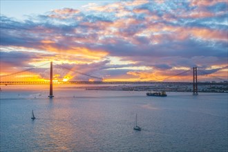 View of 25 de Abril Bridge famous tourist landmark of Lisbon over Tagus river with tourist yacht boats and cargo container ship on sunset. Lisbon