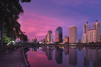 Beautiful landscape with modern high-rise buildings reflecting in calm lake waters by park and boardwalk at colorful sunset. Benjakitti Park