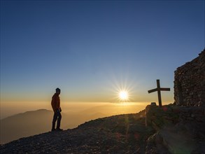 Mountaineer standing at sunset next to summit chapel Timios Stavros and summit cross of Psiloritis