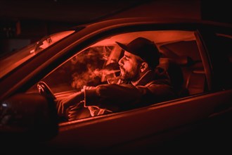 Man in a cap driving car smoking at night in a garage lit with a red light