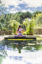 A woman holding a glass purple lotus while sitting in lotus pose on a tree trunk over the water. Vertical shot