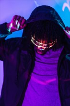 Vertical studio portrait with purple and blue neon lights of a futuristic afro man with hoodie wearing smart goggles