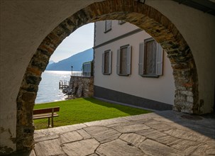 Beautiful Arch to a Park with a House on the Waterfront to Lake Lugano with Mountain in a Sunny Summer Day in Bissone