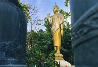 Gilded statue of standing Buddha in monk clothes and covered by golden textile shot between two bells and among trees and flowers. Right hand in adhaya mudra gesture. Selective focus
