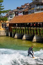 Surfer Surfing the Wave on River Aare in City of Thun from Untere Schleuse Wooden Bridge in a Sunny Summer Day in Thun