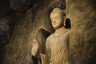 Statue of standing Buddha in monk clothes carved in limestone cave and lit by light. Right hand in adhay mudra gesture. Religion