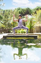 A woman practicing the yoga pose Goddess with Cactus Arms on a tree trunk over a pool