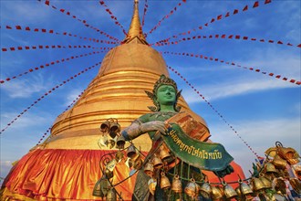 Statue of green-skinned Thai angel holding roll with message in Thai stand in front of big golden stupa in sunlight and at blue sky with clouds in Buddhist temple. Decorations with bells