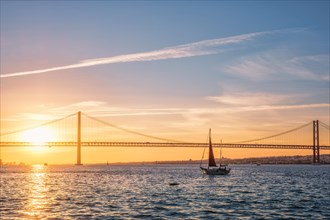 View of 25 de Abril Bridge famous tourist landmark of Lisbon connecting Lisboa and Almada over Tagus river with tourist yacht silhouette at sunset. Lisbon