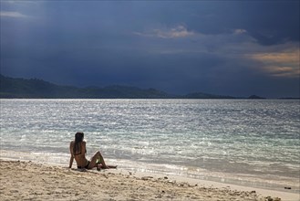 Female tourist in bikini sitting on tropical beach of the islet Gili Kedis and watching approaching thunderstorm over the island Lombok