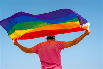 A gay person with the LGBT flag on his back moving with the wind with the sky in the background