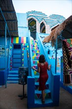A young woman in a red dress climbing some colorful stairs that go up to a beautiful terrace of a traditional blue house in a Nubian village next to the Nile river and near the city Aswan. Egypt