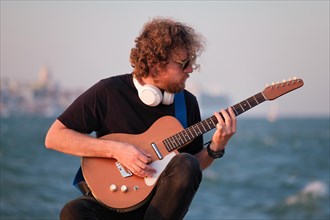 Hipster street musician in black playing electric guitar in the street on sunset on embankment
