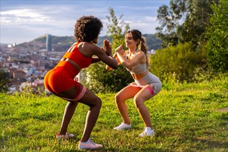Caucasian blonde girl and dark-skinned girl with afro hair doing squat exercises in a park with the city in the background. Healthy life