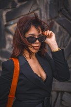 Brunette girl in the city with sunglasses and red bag on a stone wall looking at the camera