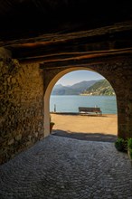 Old Beautiful Street Tunnel with Arch and a Bench from Brusino Arsizio on the Waterfront in a Sunny Summer Day and with Lake Lugano and Mountain View over Morcote