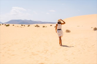 A young Caucasian tourist in a white dress and a hat walking through the dunes of the Corralejo Natural Park