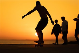 Silhouette of a man skateboarding at sunset around other people. Mid shot