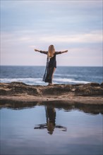 Summer lifestyle with a young brunette Caucasian woman in a long black transparent dress on some rocks near the sea on a summer afternoon. Raising arms and looking out to sea
