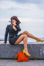 Fashion pose. Brunette girl sitting on the beach with a red bag and yellow shoes looking to the left