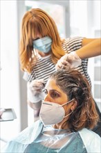 Blonde hairdresser with face mask giving the dark tint to the client at the hairdresser. Safety measures for hairdressers in the Covid-19 pandemic. New normal