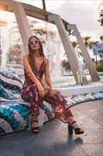 Portrait of a young brunette in a maroon floral dress enjoying the summer in the golden hour in a city fountain