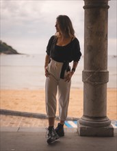 Lifestyle in the city with a blonde girl in white pants and a leather jacket near the beach. Photos next to a column in the shallows of the beach looking to the left