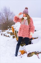 Blonde girl in pink fur jacket and purple hat in the snow looking at the horizon. Next to some trees cut with ice
