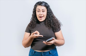 Amazed girl using and pointing cell phone looking at camera isolated. Portrait of surprised latin woman holding cell phone and looking at camera