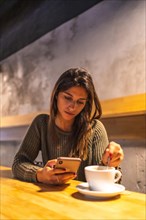 A young Caucasian brunette entrepreneur working from a coffee shop. Sending a message with the mobile phone and having a coffee