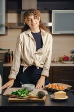 Positive young caucasian woman smiling in the kitchen standing next to table with ingredients for scrambled eggs