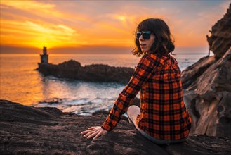 A young girl sitting with sunglasses in the orange sunset of the Pasajes San Juan lighthouse