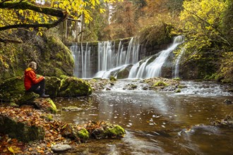 The Geratser waterfall in autumn. A hiker sits on a rock. Moss-covered rocks on the sides