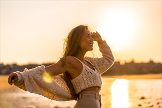 Summer lifestyle. A young blonde Caucasian woman in a white short wool sweater on a beach sunset. With open arms by the sea