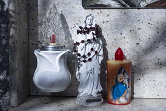 Kitschy electric battery-powered candles next to a small rosary-wrapped figure of Mary on an urn grave in a cemetery