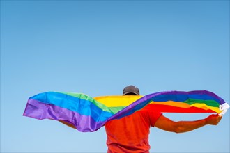 A gay person with a pink t-shirt and black cap with the LGBT flag on his back moving with the wind with the sky in the background