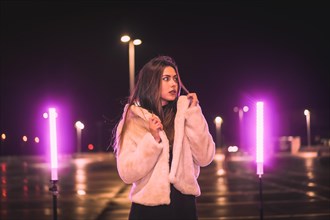 Young Caucasian brunette with wool jacket in an empty car park illuminated with pink light. Night urban winter session in the city