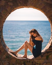 A young pretty red-haired Caucasian girl smiling sitting in a blue dress by the sea