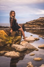 Summer lifestyle with a young brunette Caucasian woman in a long black transparent dress on some rocks near the sea on a summer afternoon. Sitting on top of a rock in a natural pool