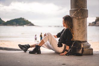 Lifestyle in the city with a blonde girl in white pants and a leather jacket near the beach. Sitting on the shallows of a beach on a beautiful sunset