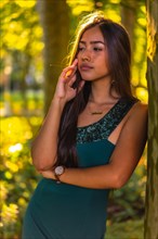 A young brunette Latina with long straight hair leaning against a tree in a green dress. Portrait with a sweet and sensual look