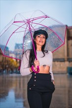 Posing of a young brunette Latina with a leather cap in the rain of the city with a transparent umbrella