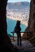 A young hiker wearing a hat in the tunnel of the Penon de Ifach Natural Park with the city of Calpe in the background