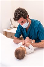 Frontal photo of a young doctor with facial mask examining a cute newborn baby in a clinic